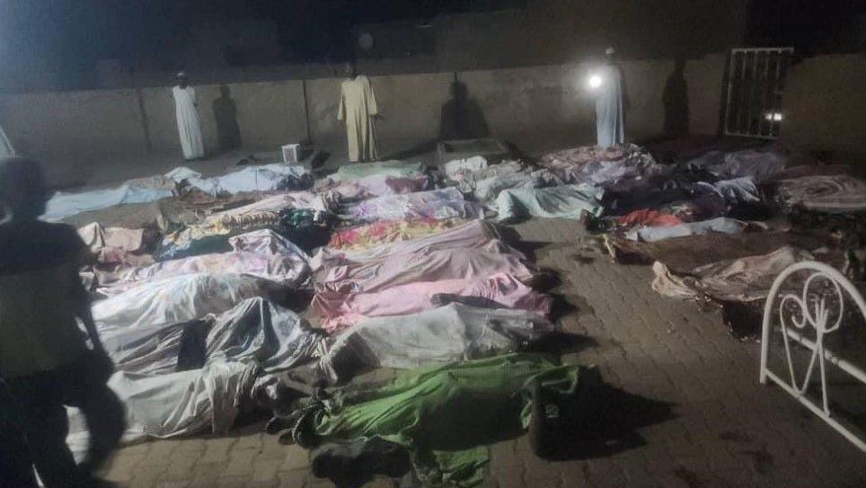 Dead bodies lay out in the mosque courtyard. They are covered by cloth as men stand around. 