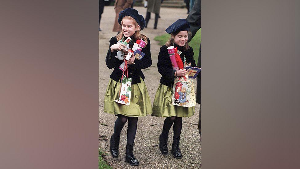 Young girls wearing matching outfits of beret-style hats, gold skirts and velvet jackets. They are walking outside.
