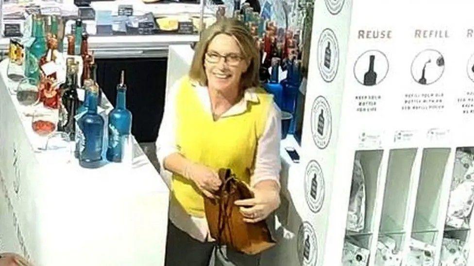 Gaynor Lord on CCTV dressed in a white shirt, with a yellow tank top and is carrying a brown bag. She has blonde hair and glasses and is pictured at work at a gin counter.