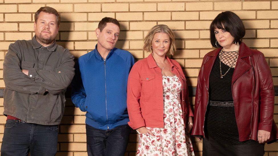 Four members of the cast of Gavin and Stacey reunite for an official cast photo: Smithy, Gavin, Stacey and Nessa. They are stood against a brick wall. Smithy (played by James Corden) is wearing a grey jacket and blue jeans and has his arms crossed, Gavin (played by Matthew Horne) is next to him, wearing a blue jacket with his hands in the pockets and blue jeans. Next to him is Stacey (played by Joanna Page), she is wearing brown cowboy boots, a white floral dress and a pink jacket, and she is stood next to Nessa (played by Ruth Jones), who is wearing a burgundy leather jacket and a black dress with long black boots.