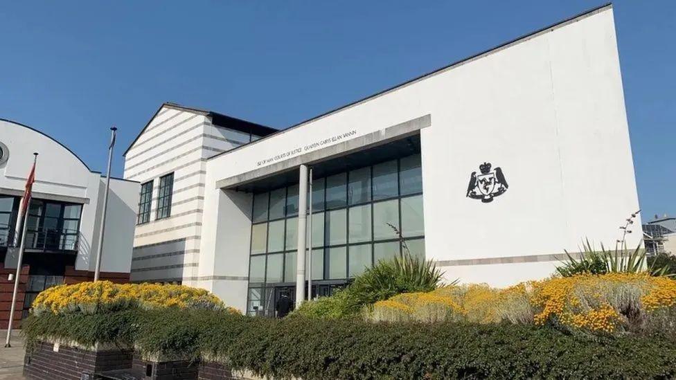 The exterior of the white courts of justice building, a large white glass-fronted building with the Isle of Man Courts of Justice written above the entrance and the government crest to the right.