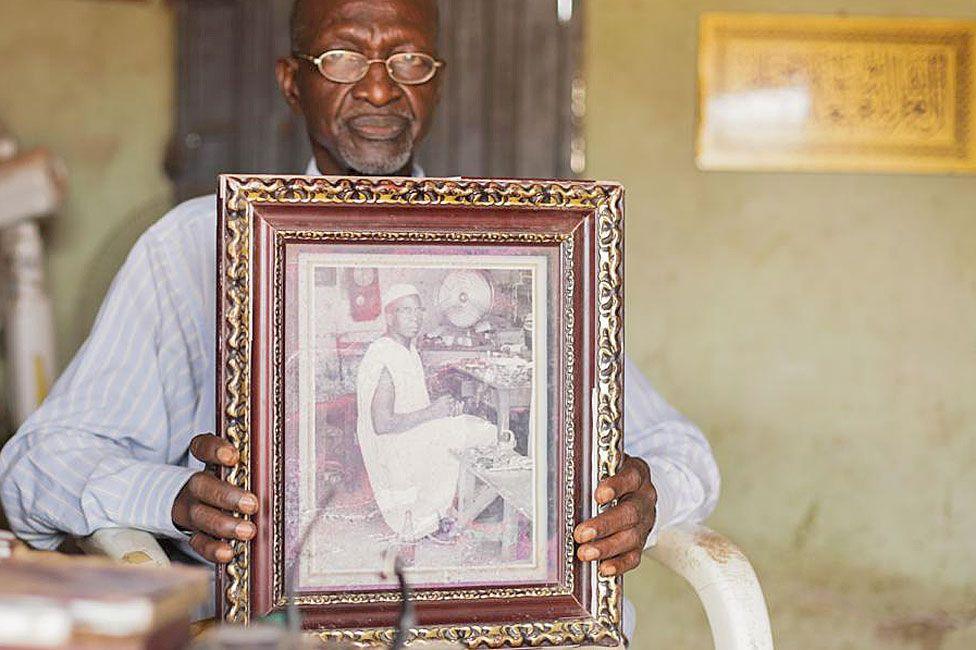 Bala Muhammad, wearing wire-rimmed glasses and a long-sleeved pin-stripped blue shirt, sits in a white-painted wooden chair holding up a wide-wooden framed black-and-white photograph of his father, who is pictured looking at the camera as he sits at his workshop bench. He is wearing a sleeveless boubou. A desk fan can be seen in the background and a pendulum clock hangs on the wall