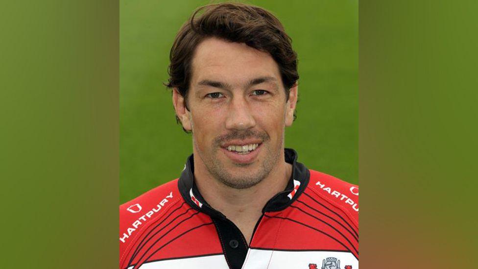 A head and shoulders shot of Tom Voyce in a Gloucester rugby kit. It is a red and white hooped shirt with the logo of the company Jewson across the chest.