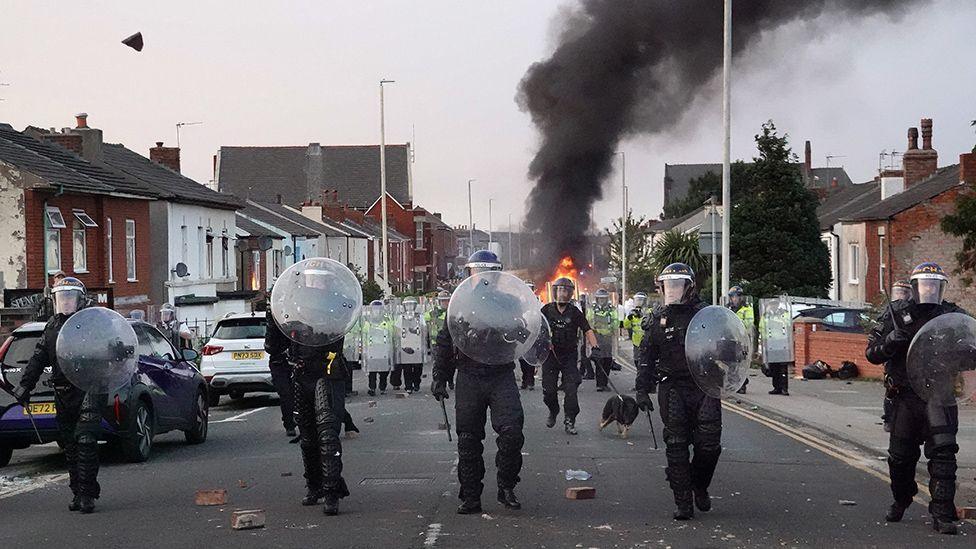 Four riot police, holding shields walk down a residential street in Southport on a summer's evening. Bricks are strewn over the road, and in the background, a fire is burning. Behind the four police in the foreground, a line of police with longer shields follow them. 