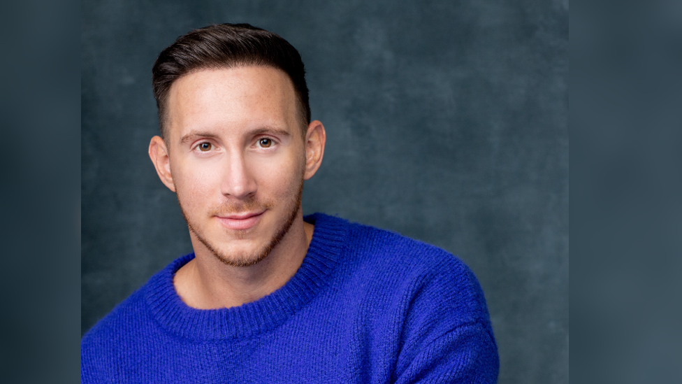 A headshot of Rhys Batten, looking at the camera in front of a dark background. He is wearing a blue jumper.