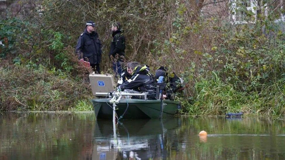 Divers and police officers in a small boat filled with equipment near a bank on the River Wensum. Two men are standing on the bank, with a building visible behind trees and bushes.