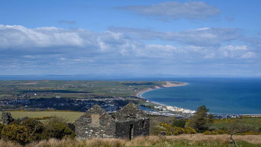 The view from the Gooseneck looking over Ramsey Bay. In the foreground there are the ruins of an old cottage, and beyond that a town that sits by sea. There is a collection of buildings on the seafront backed by lots of green fields the further inland.