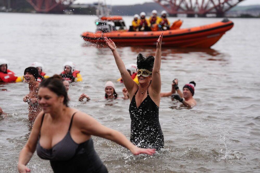 People in swimming costumes and hats race out of the water in the Forth of Firth. A lifeboat can be seen in the background.