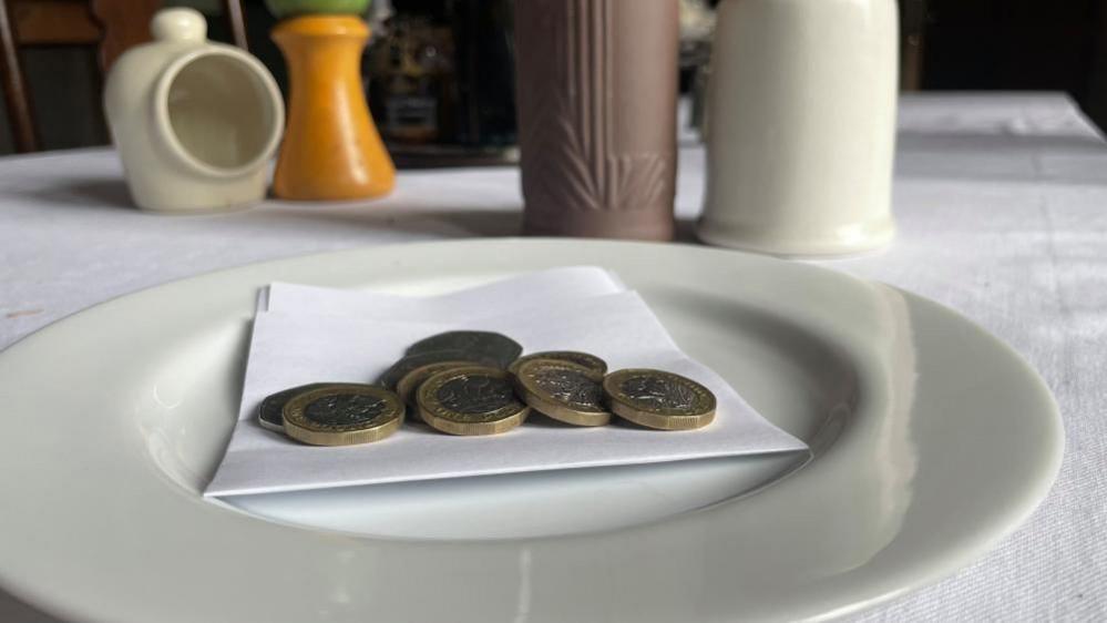 Coins on a piece of white paper, on a plate at a table set with bottles and condiments - a picture representing a customer tip.