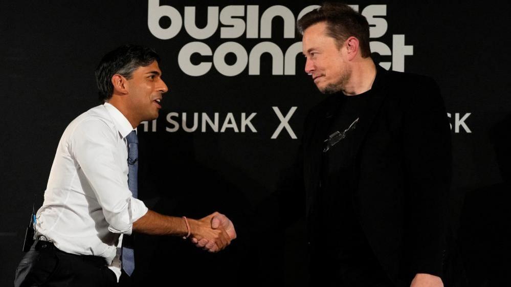 A man in a white shirt and tie shakes hands with a man in a dark top on a conference stage