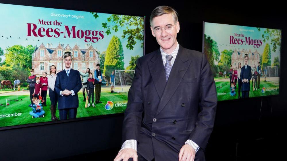 Jacob, smiling, wearing a blue jacket and tie, sitting in front of some posters for the TV show Meet the Rees-Moggs