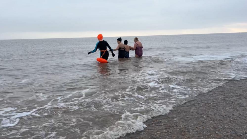 Pam Spychal, dressed in an orange hat and black swim suit, and orange floatation marker, with three friends entering the sea at Great Yarmouth beach