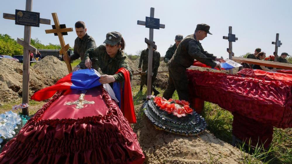 Several Russian soldiers pin flags to coffins of soldiers killed in Luhansk, Russia-controlled territory, surrounded by crosses