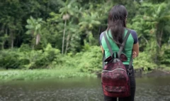 Menina de costas com mochila e uniforme escolar observa o rio, com floresta ao fundo. 