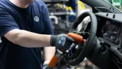 Um homem montando o volante de um carro em um fábrica com uma ferramenta laranja