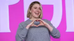 Greta Gerwig sorrindo e fazendo coração com as mãos