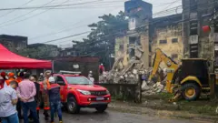 Imagem mostra bombeiros e retroescavadeira em frente a prédio que desabou