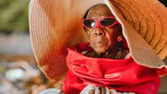 Margret Chola looking glamourous in a oversized wicker hat, red leather jacket, gold jewellery and red sunglasses