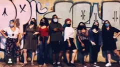 Os oito membros do coletivo PerifAnálise posam para foto, usando máscaras de proteção contra o coronavírus, e em frente a um muro coberto de grafites