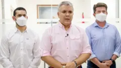 Duque em pé fazendo discurso em sala, com outros dois homens ao lado