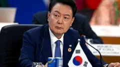 Yoon Suk Yeol sentado com um microfone e uma bandeira em miniatura da Coreia do Sul na sua frente em 19 de novembro de 2024