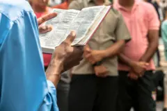 Pastor lendo a Bíblia, observado por pessoas em pé atrás