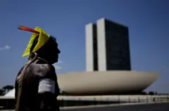 Homem indígena com cocar de penas em frente ao prédio do Congresso Nacional 