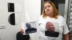 Antônia Regina segura foto do filho e usa camiseta em homenagem a Denner com a frase "Justiça pelo meu filho Denner"