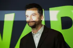 Fotografia de Wagner Moura, um homem branco de barba de terno sorrindo com uma faixa de luz sobre seus olhos em frente a um cartaz escrito "war"