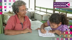 Seca na Amazônia: idosa de pele escura e cabelo curtos sentada à mesa ensinando uma criança também de pela escura, que realiza atividade escrita