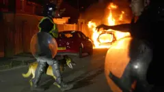 Policial passa com cão em rua com um carro em chamas ao fundo 