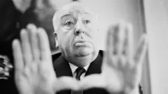 Alfred Hitchcock, um senhor de pele clara, com pouco cabelo, em uma foto em preto e branco. Ele está vestindo terno escuro e gravata e aparece com os braços estendidos e as duas mãos abertas, com a ponta de um dedão encostada na outra. 