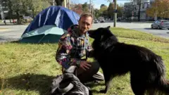 Homem agachado ao lado de cachorro, em gramado com barraca ao fundo, em zona urbana