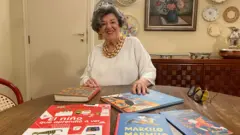 Ruth sorrindo sentada diante de mesa com vários livros