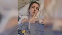 Faye Louise está deitada em uma cama de hospital, com vários fios ao redor de seu corpo, e coberta com uma bata azul.
