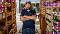 A foto mostra Ednilson, um homem de pele morena, vestindo uma camiseta polo azul-marinho, calça jeans e boné. Ele está em uma loja e olha para a câmera de braços cruzados.