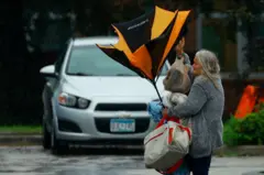 Mulher em estacionamento segurando sacolas e guarda-chuva quebrado por conta dos ventos