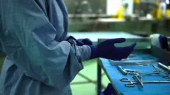 A woman wearing protective clothing holds her gloved hands out towardsw surgical instruments