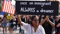 Mulher levanta bandeira dos EUA perto de cartaz que diz: 'Uma vez imigrante, sempre um sonhador'