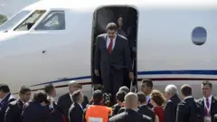 A foto mostra Maduro, um homem de pele clara, cabelo e bigode pretos, vestindo um terno escuro sobre camisa branca e gravata vermelha, descendo da escada de um avião. 