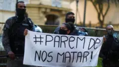 Fotografia colorida mostra mulher jovem negra segurando cartaz "parem de nos matar"  