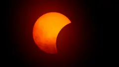 Foto colorida mostra o início do eclipse, com o Sol na parte de trás, laranja, e a lua o encobrindo como uma sombra