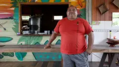 Roberto Brito, um homem corpulento vestindo camiseta vermelha, posa em frente a uma mesa de madeira e a uma construção também em madeira, pintada com um desenho colorido de folhas amazônicas