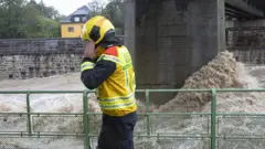 Foto de um agente de segurança, com uma roupa amarela e preta e capacete amarelho em uma ponte sobre um rio com águas agitadas 