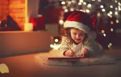 Menina com gorro de Papai Noel deitada no chão escrevendo carta com árvore de Natal iluminada ao fundo