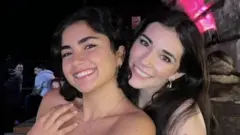 Duas garotas sorrindo para a câmera. À esquerda, a garota tem cabelo escuro na altura dos ombros. Ela está usando uma blusa jeans azul e um colar prateado. A garota à direita tem cabelo escuro e longo. Ela está usando uma blusa jeans azul e brincos prateados. A foto foi tirada em um bar com longas mesas de madeira ao fundo.