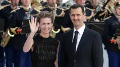 Ex-presidente sírio Bashar al-Assad (à direita) e sua esposa Asma (à esquerda) durante um jantar na cúpula UE-Mediterrâneo em Paris em 13 de julho de 2008. Asma acena para o público. Atrás deles estão membros da Banda Presidencial da França.