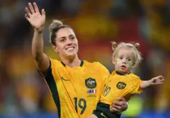 Katrina Gorry porte sa fille Harper, âgée de deux ans, tout en portant des kits de football assortis, alors qu'elles saluent la foule.