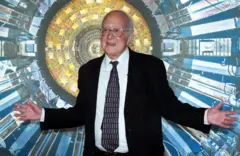 Peter Higgs em frente a painel com imagem do que parece ser um equipamento