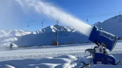 Canhão dispara neve artificial, com teleférico e pessoa com snowboard atrás